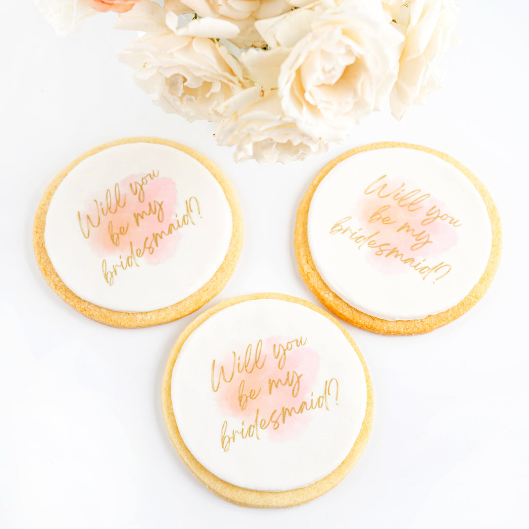 Three cookies with 'Will you be my bridesmaid?' text on a white background with flowers.