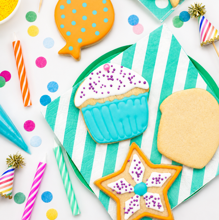 A styled flat-lay of decorated sugar cookies on striped paper napkins with party props, including a balloon-shaped cookie, a cupcake-shaped cookie, a star-shaped cookie, candles, confetti, and party hats on a white background.