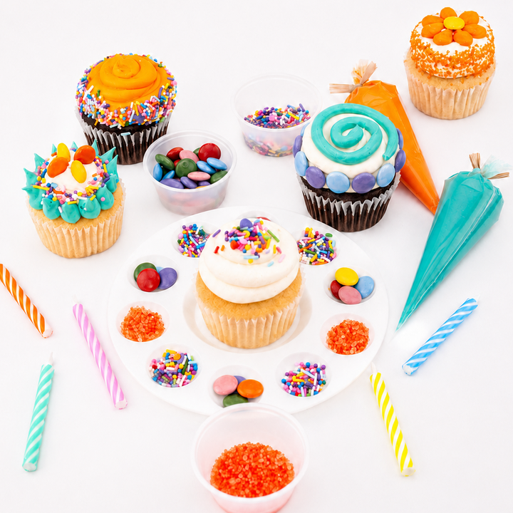 A styled flat-lay of assorted cupcakes with colorful frosting, sprinkles, candy, piping bags, and striped candles on a white background.