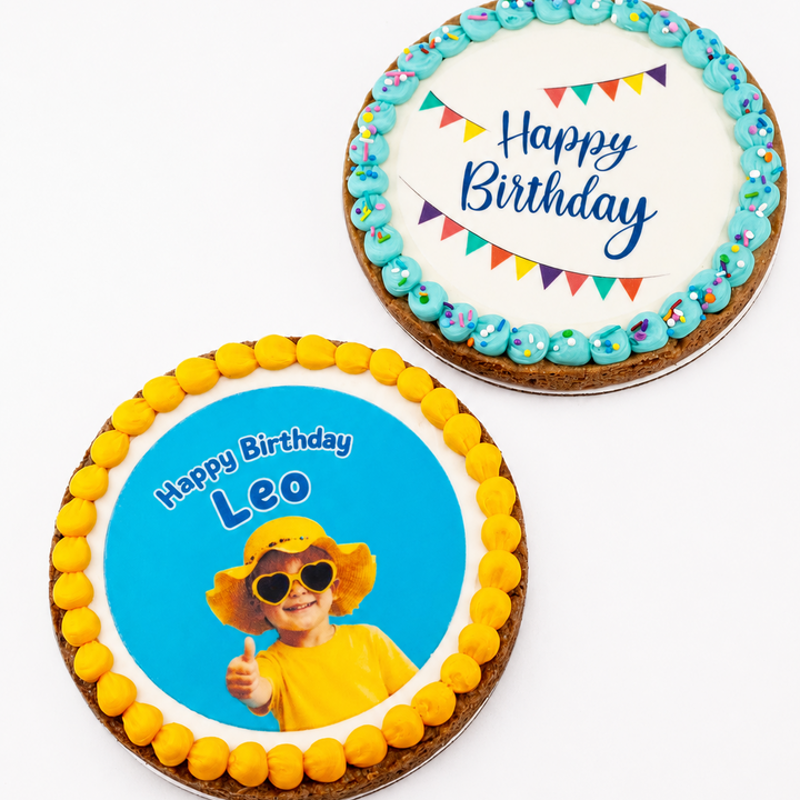 Two round birthday cakes on a white background. One cake has a white icing top with blue 'Happy Birthday' script, colorful bunting, and blue iced cookies around the edge.