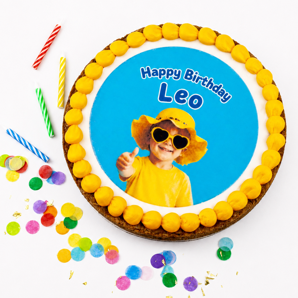 A round birthday cake topped with yellow frosting and a custom printed photo of a child wearing sunglasses, with the text 'Happy Birthday Leo'. The cake is decorated with colorful candy canes and scattered confetti on a white background.