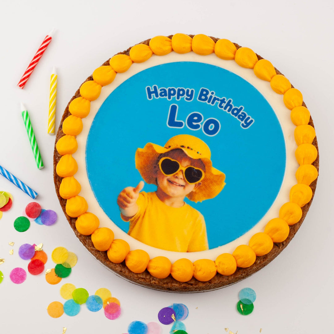 A round birthday cake with a custom printed photo of a child in a yellow hat and sunglasses on a bright blue background, surrounded by a border of yellow frosting. The cake is decorated with colorful candle-shaped picks and scattered confetti on a white background.
