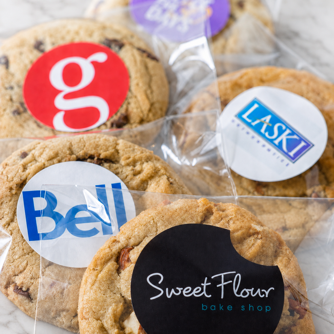 Several chocolate chip cookies in clear plastic bags with round printed stickers, including visible text such as Bell, LASKI, and Sweet Flour bake shop.