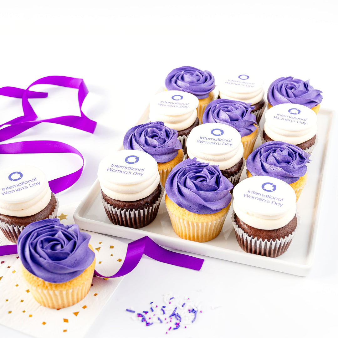 International Women's Day Cupcakes with purple and white icing on a white tray with a white background