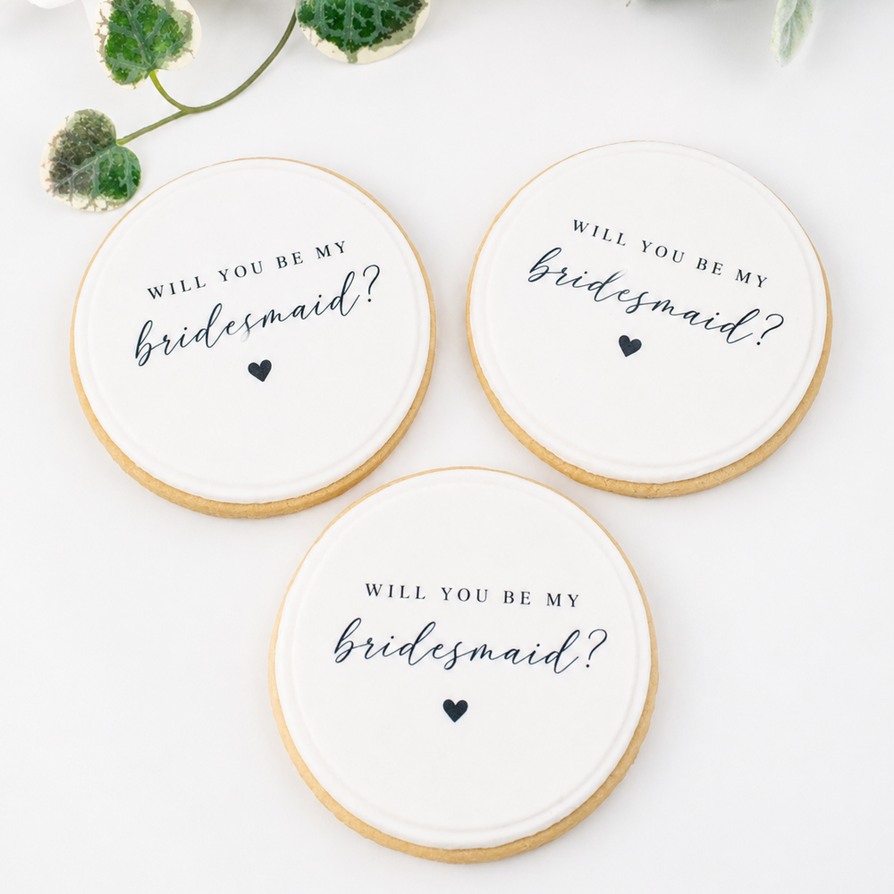 Three round white cookies with 'WILL YOU BE MY bridesmaid?' in black script and a small heart, arranged on a white surface with floral decorations in the corner.