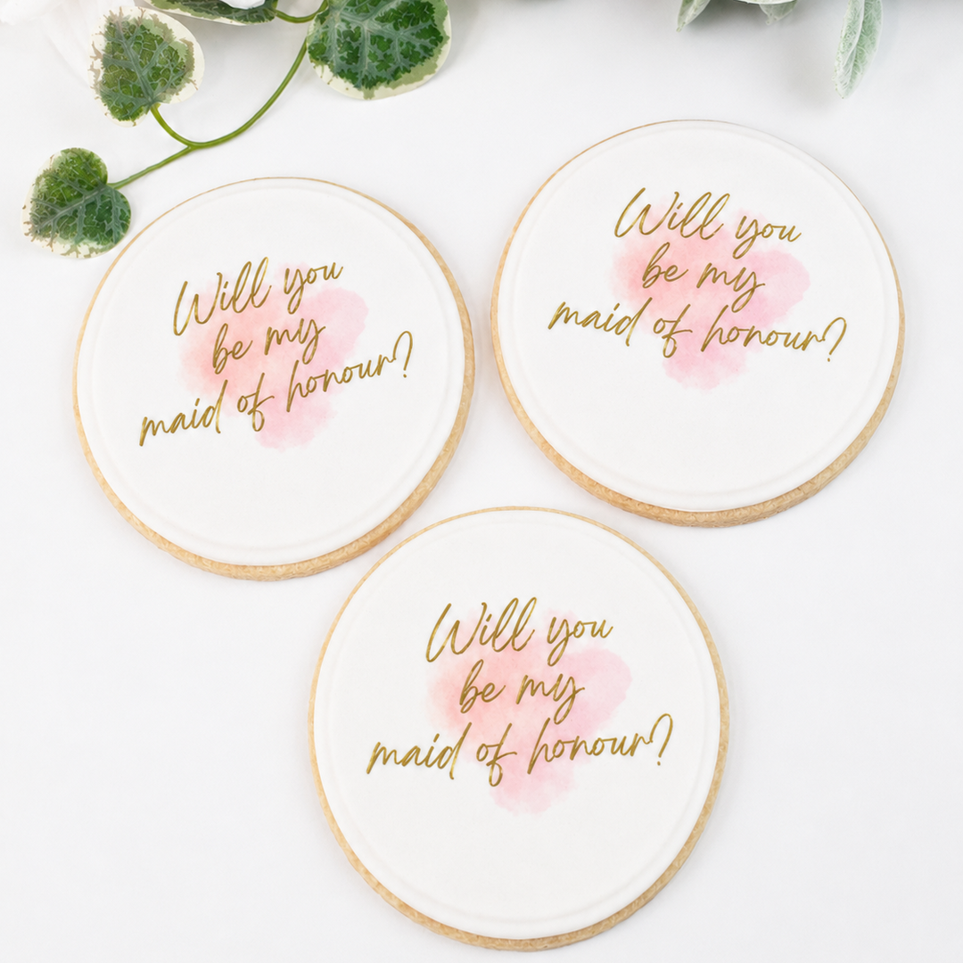 Three round white wedding cookies with gold foil lettering and pink watercolor accents, arranged on a white surface with decorative white and pink flowers at the top.