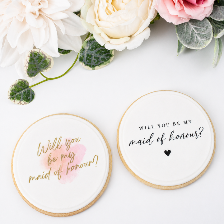 Two round white bridal shower cookies with gold and black lettering, styled with white and pink flowers on a white background.