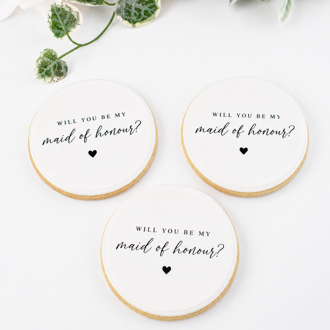 Three round white wedding cookies with gold edges, printed with the text 'WILL YOU BE MY maid of honour?' in black script and a small heart, arranged on a white surface with floral decorations in the corner.