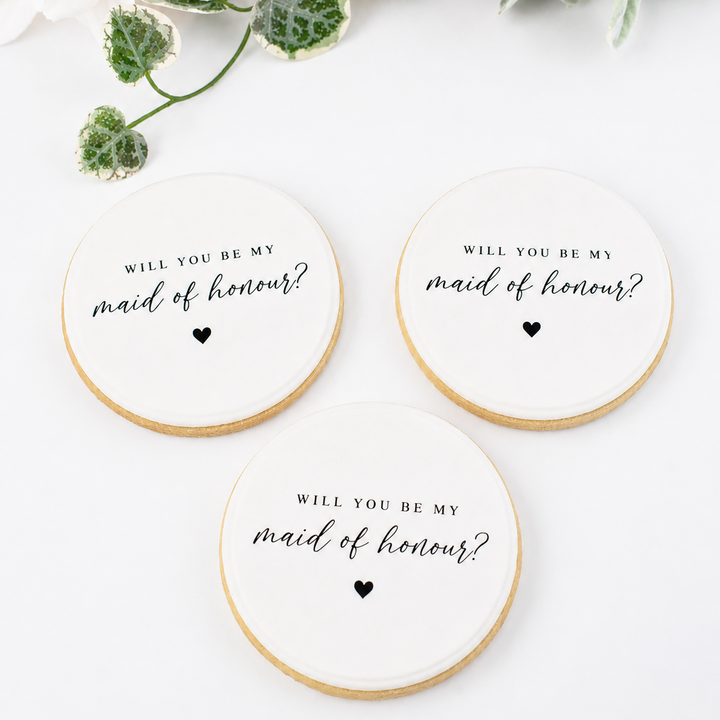 Three round white wedding cookies with gold edges, printed with the text 'WILL YOU BE MY maid of honour?' in black script and a small heart, arranged on a white surface with floral decorations in the corner.